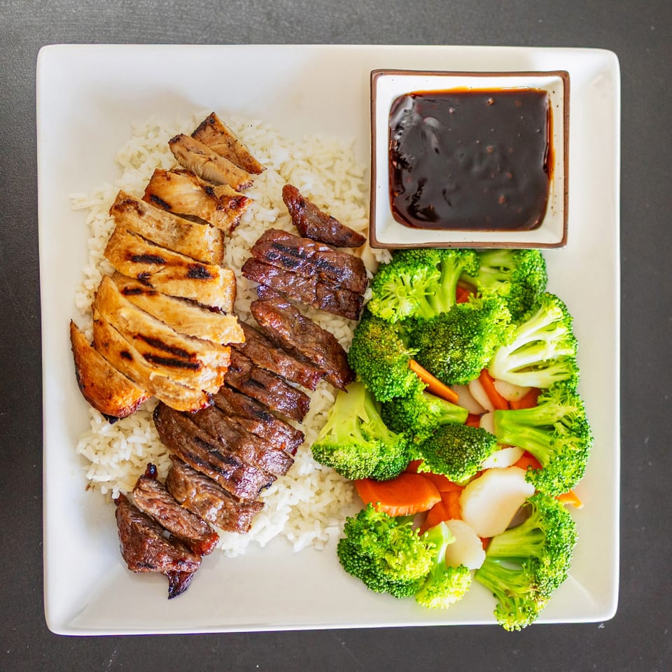 Grilled chicken and beef with rice, steamed broccoli, carrots, and a side of sauce.